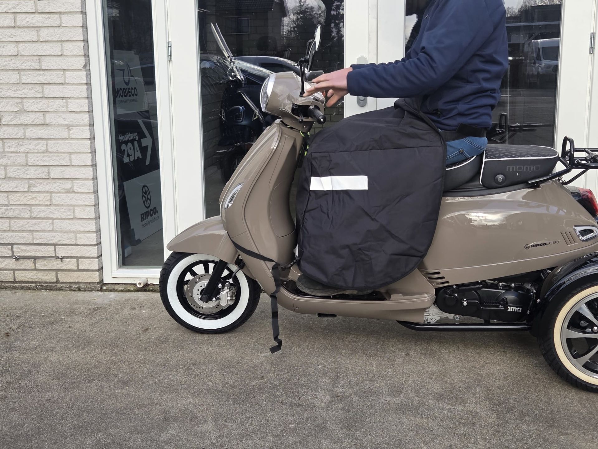 Persoon op een beige scooter met beenbeschermers. Witte banden, reflecterende strip op de beschermer, geparkeerd voor een gebouw.