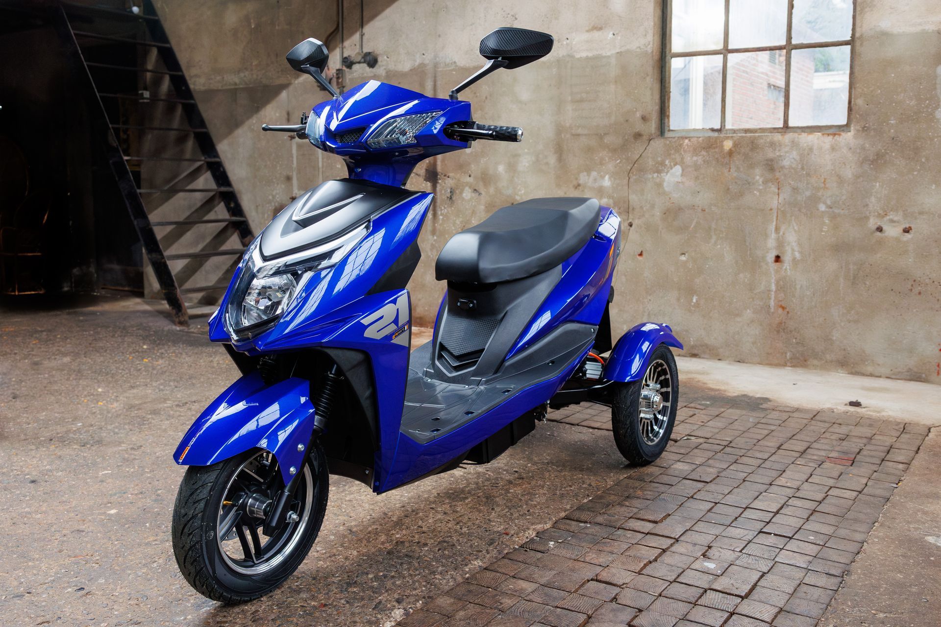 Een blauwe elektrische driewieler staat binnen geparkeerd, vlakbij een betonnen muur en een metalen trap.