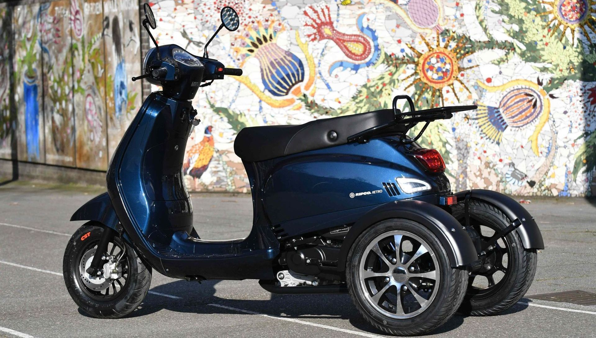 Een blauwe driewielige scooter staat geparkeerd voor een kleurrijke muurschildering.