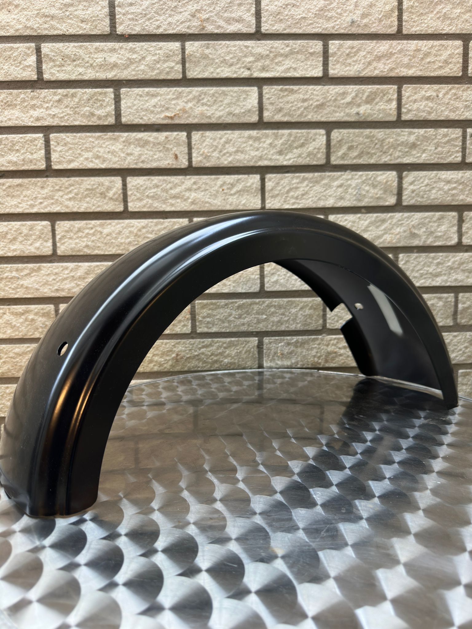 Zwarte, gebogen spatbordrand op een metalen ondergrond tegen een bakstenen muur.