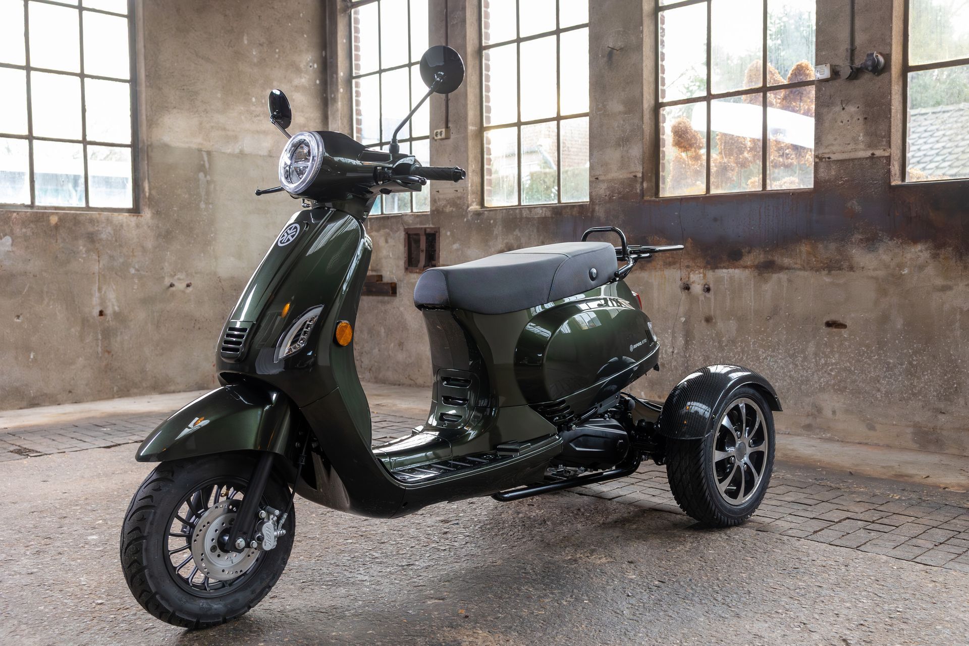 Een blauwe driewielige scooter staat geparkeerd in een industriële ruimte met grote ramen.