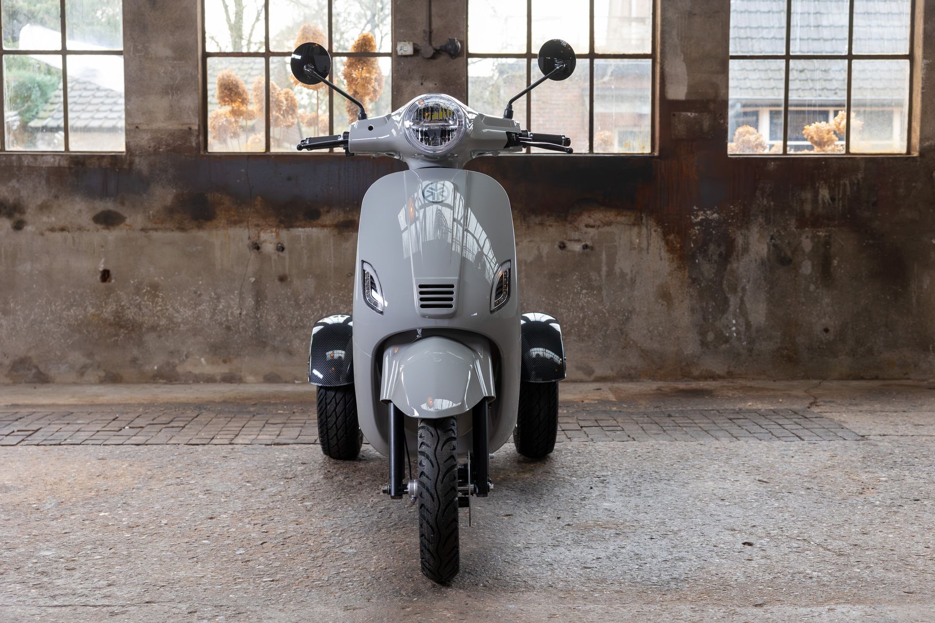 Een grijze driewielige scooter staat geparkeerd in een rustieke, industriële ruimte voor grote ramen.