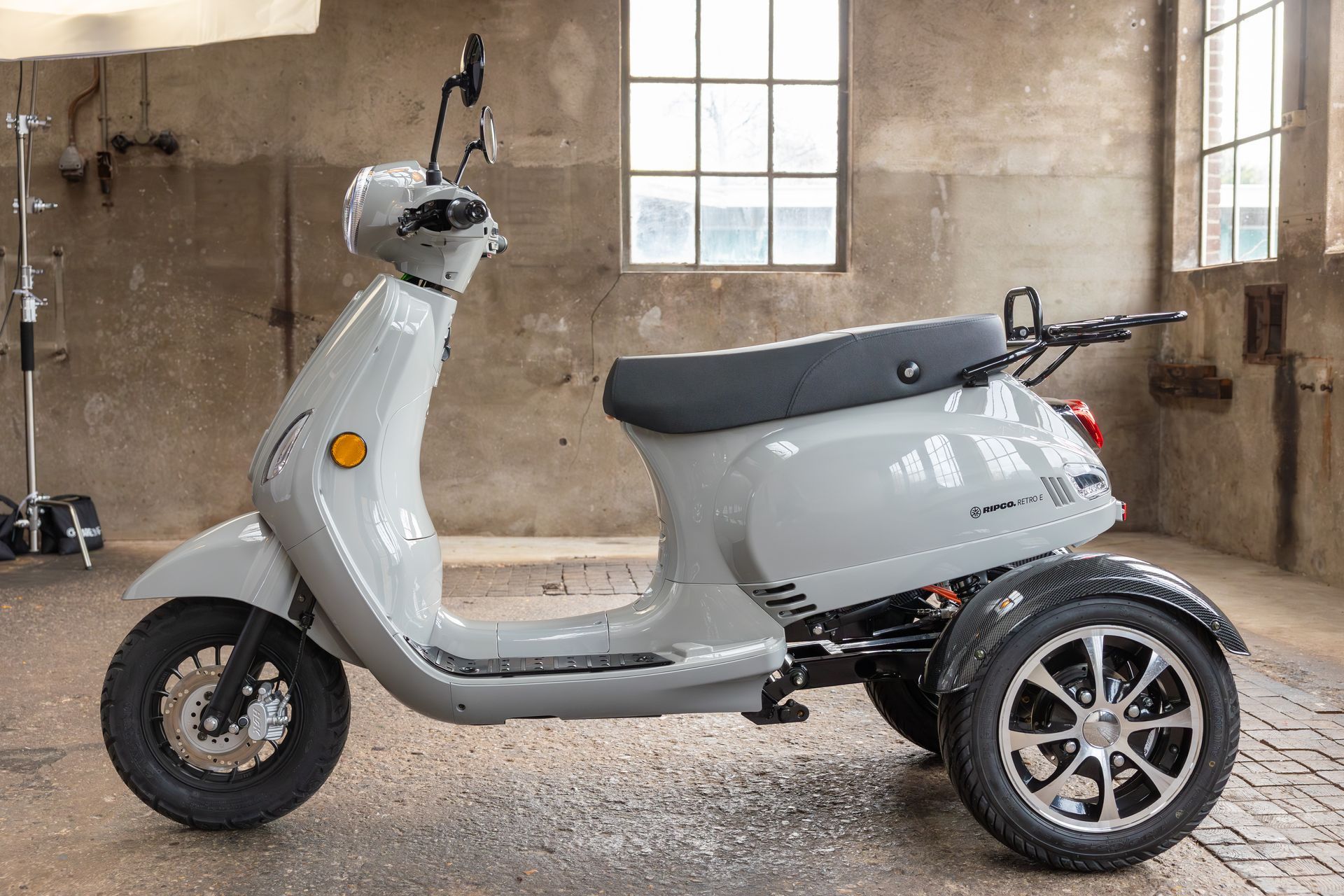 Een grijze driewielige scooter staat geparkeerd in een rustieke, industriële ruimte voor grote ramen.