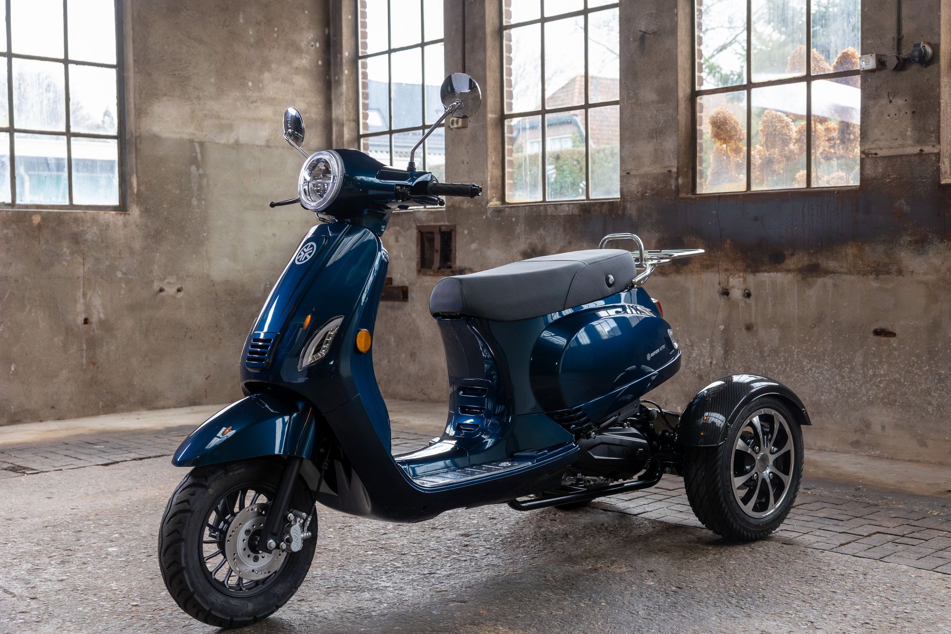 Een blauwe driewielige scooter staat geparkeerd in een industriële ruimte met grote ramen.