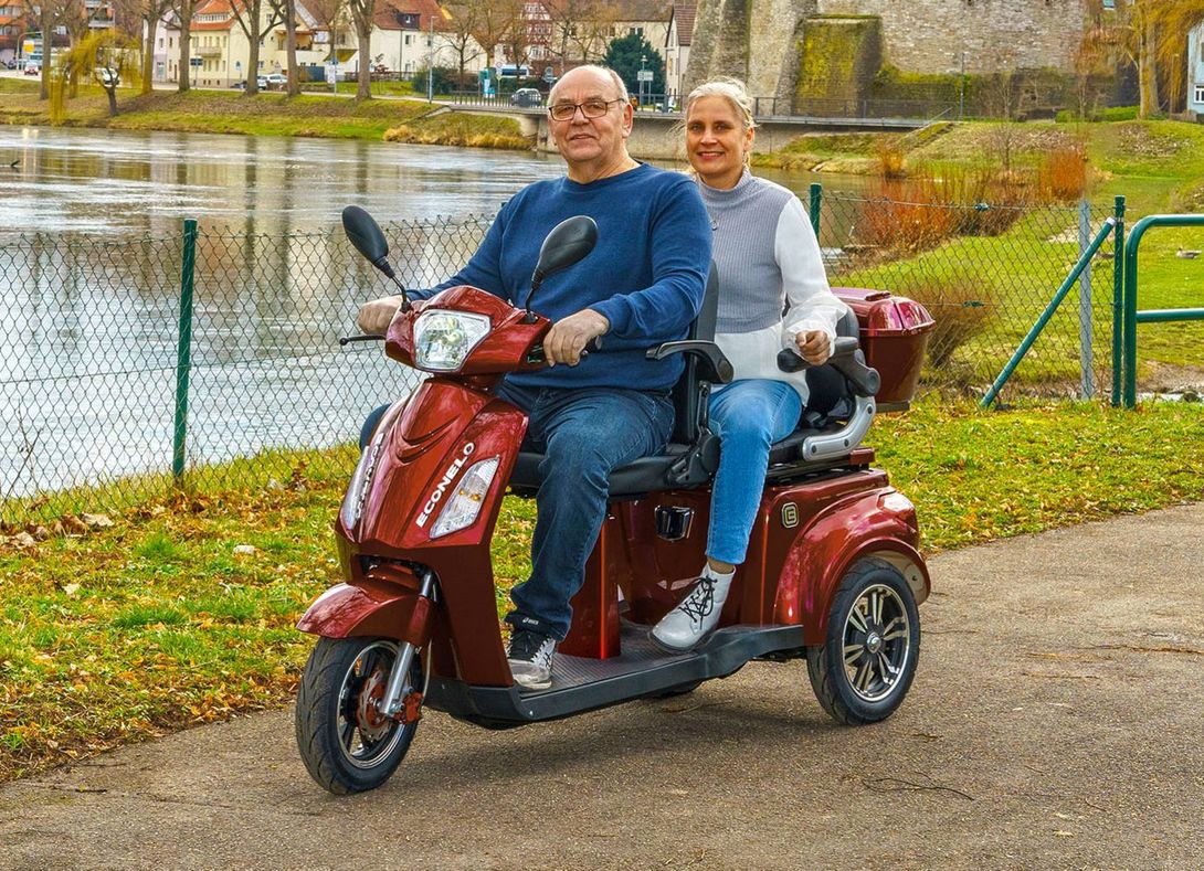 Een stel op een rode scootmobiel zit buiten, naast een vijver. Ze glimlachen.