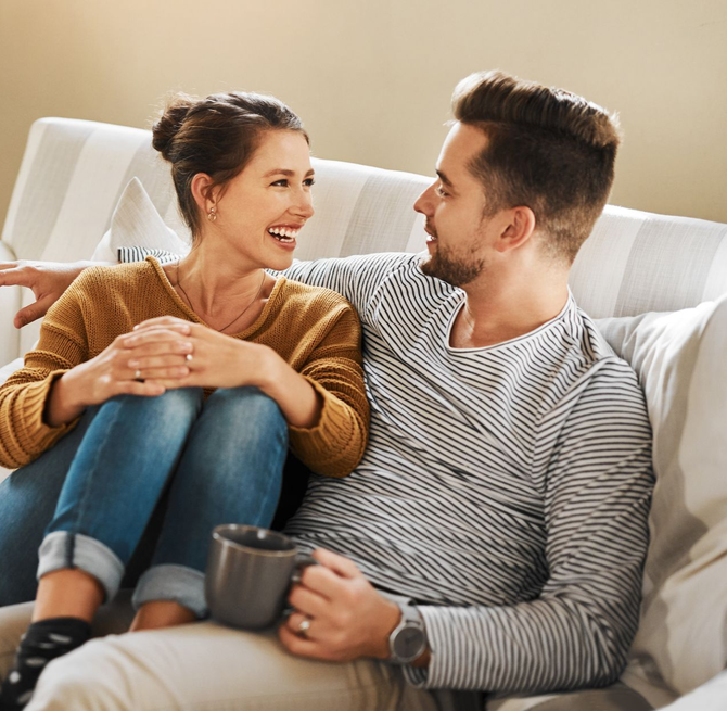 Two people smiling on a couch, talking and holding mugs in a cozy living room