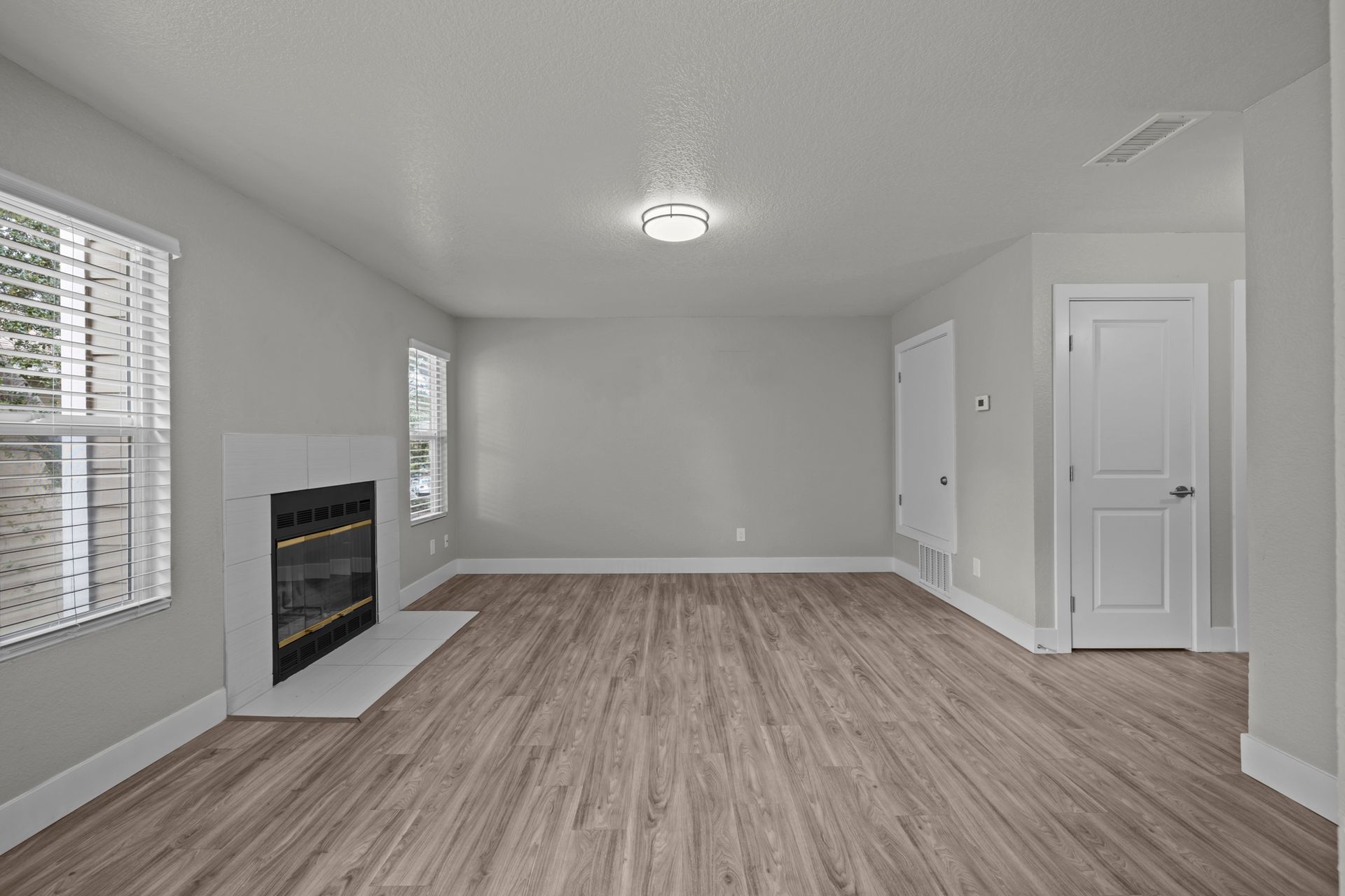 Empty modern living room with gray walls, light wood floors, fireplace, and windows with blinds
