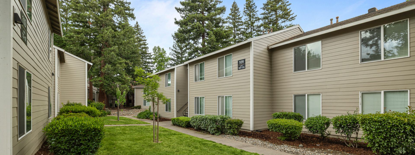 Apartment courtyard with beige buildings, green lawn, shrubs, and tall trees under a clear sky