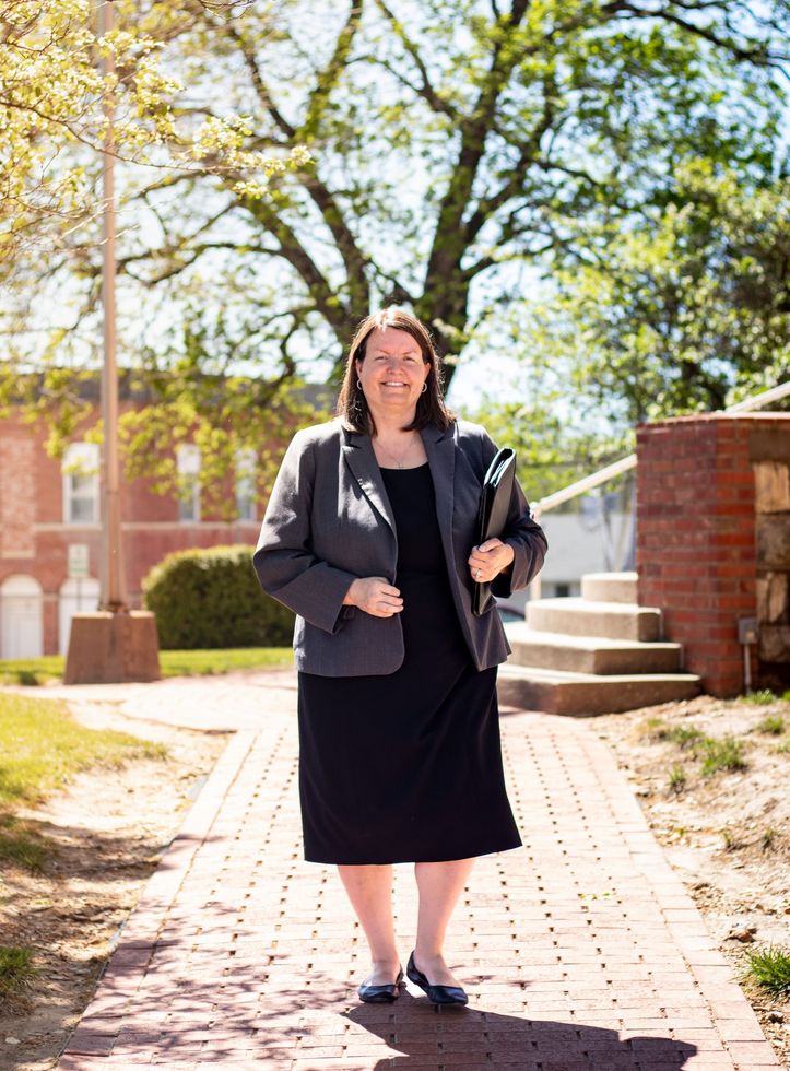 Woman in a dark outfit standing on a brick path outdoors by brick buildings and trees, holding a diploma folder.