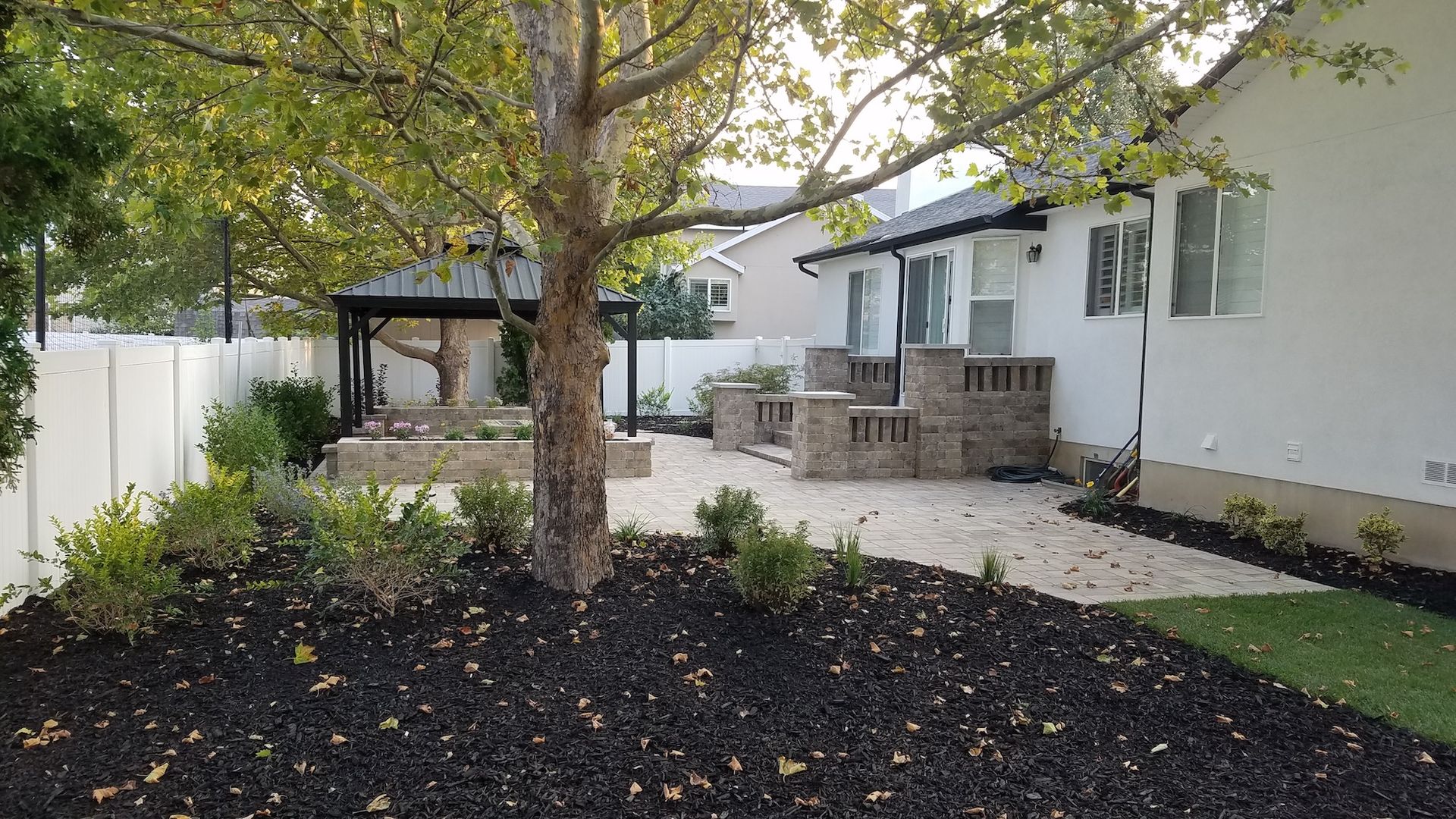 Backyard with tree, stone patio, white fence, and landscaping.