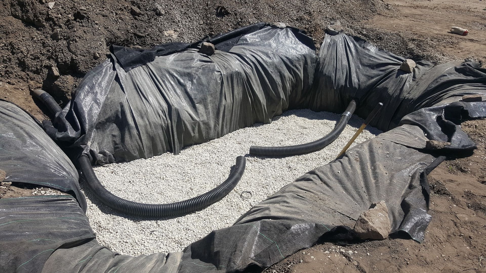 Black liner in a dug hole, filled with gravel and a curved black pipe.
