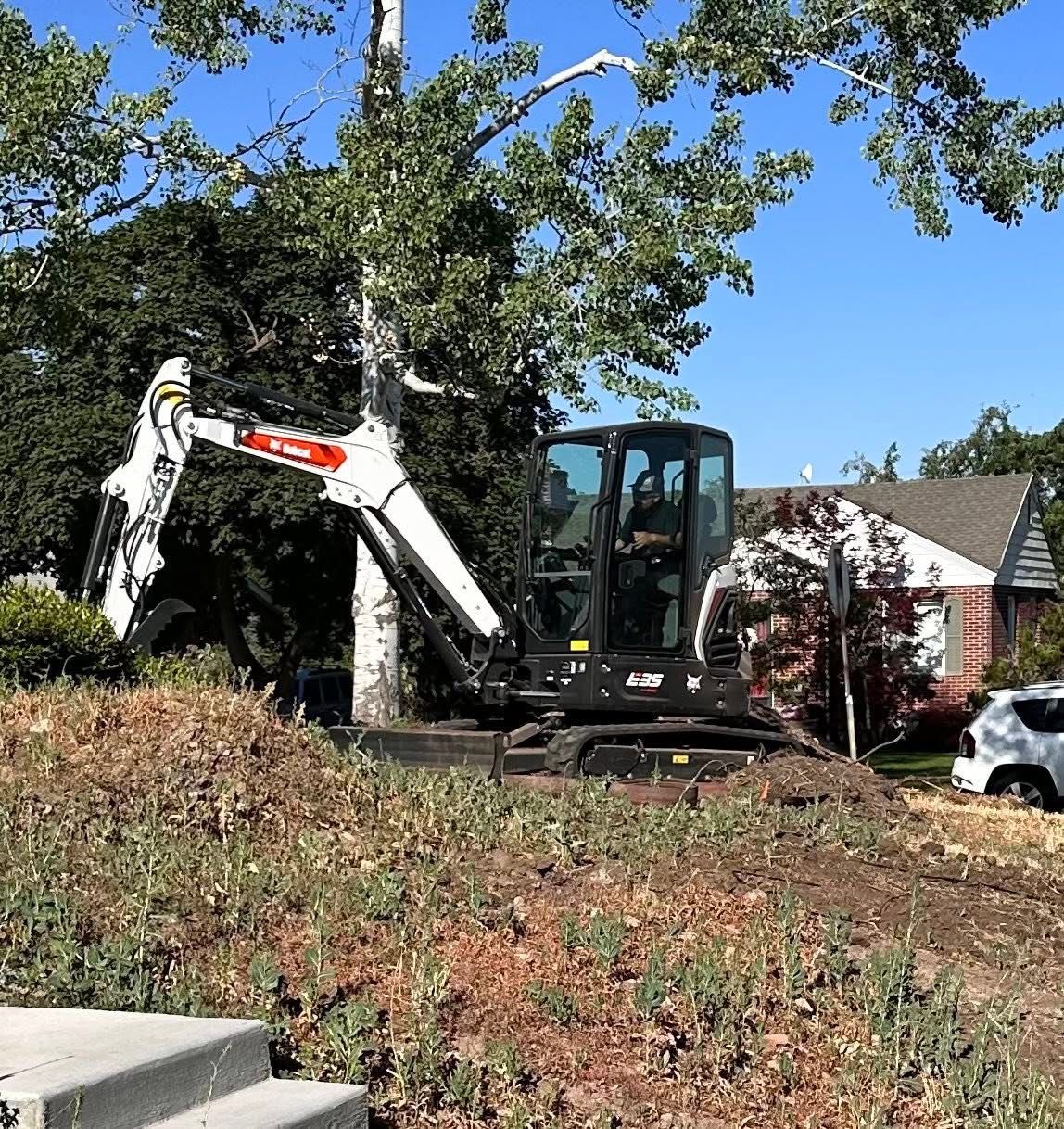 Bobcat excavator working near a tree in front of a house on a sunny day.