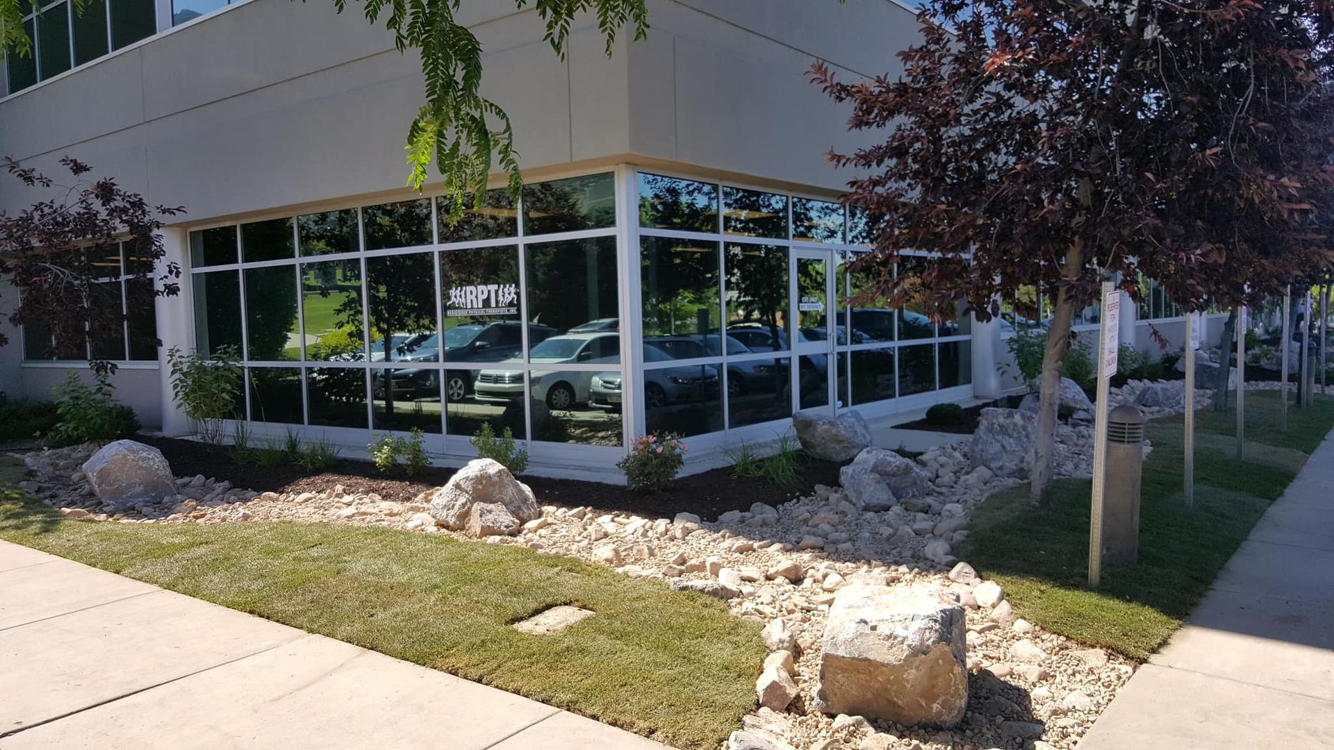 Corner of a modern building with large windows reflecting cars; landscaped with rocks and trees.