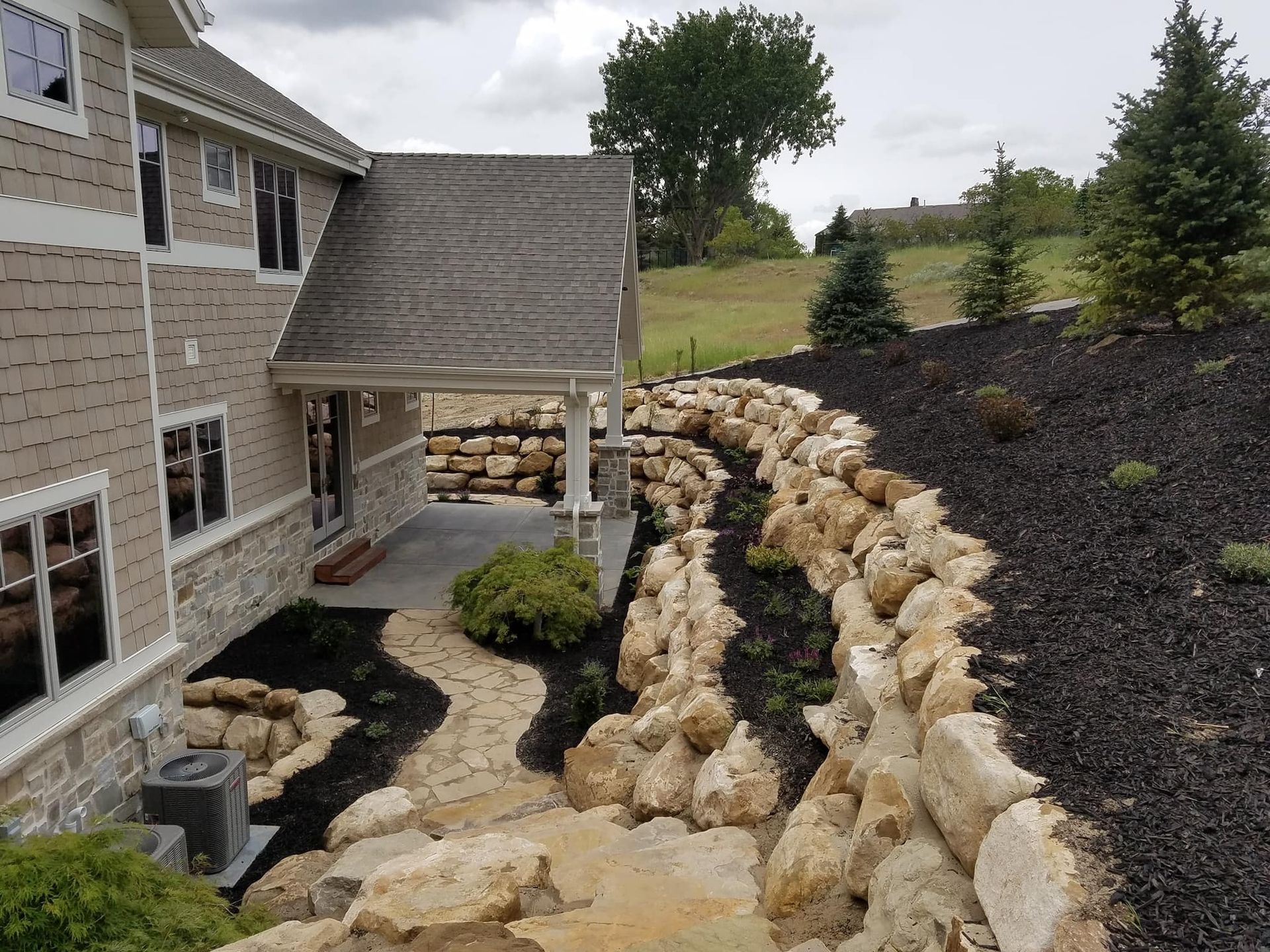 Exterior view of a house with a stone retaining wall and winding stone pathway. Black mulch and landscaping present.