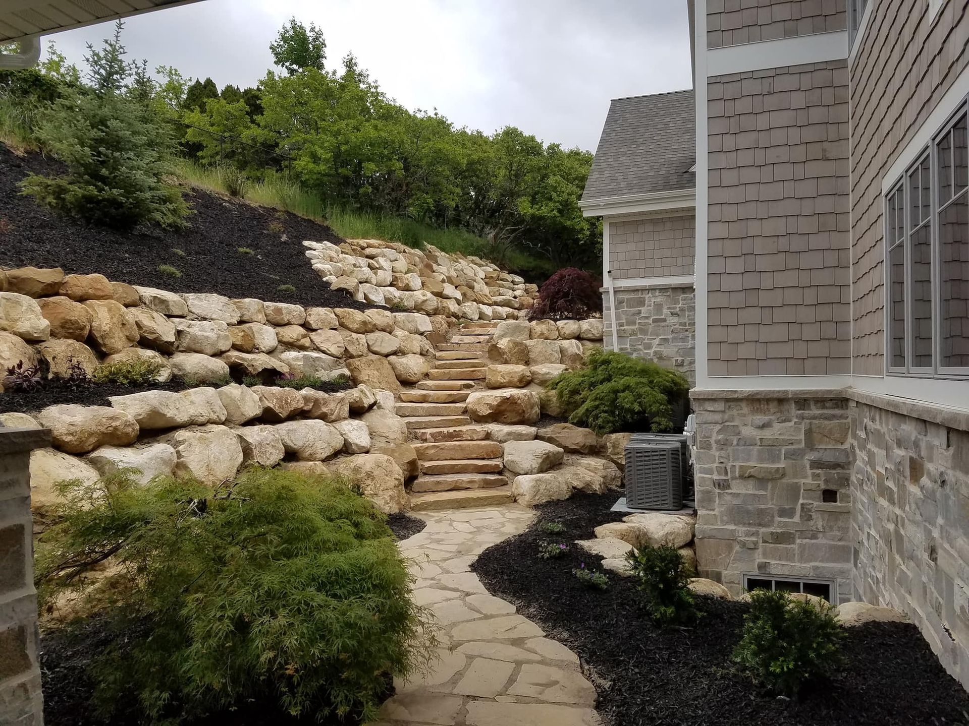 Stone staircase and path leading up a hillside with rock retaining walls and landscaping, next to a house.