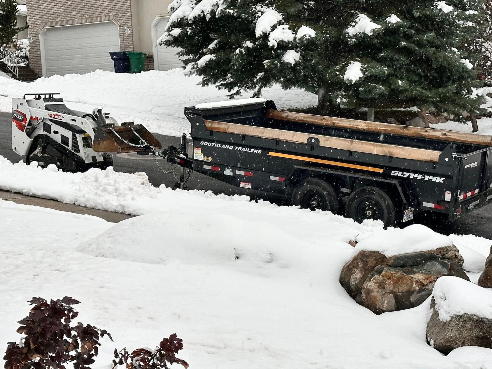 A Bobcat skid steer loading snow into a black trailer on a snow-covered residential street.