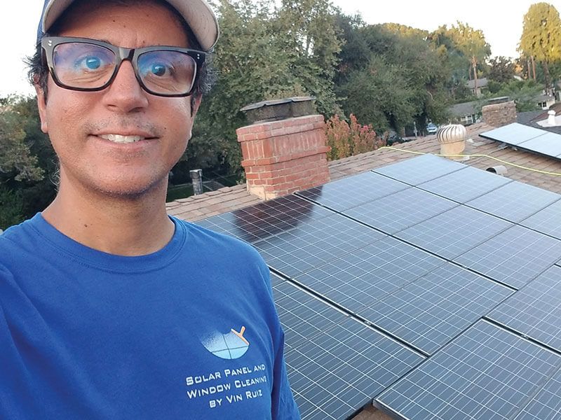 A professional cleaner wears a blue shirt and smiles in a selfie on a rooftop next to installed solar panels.