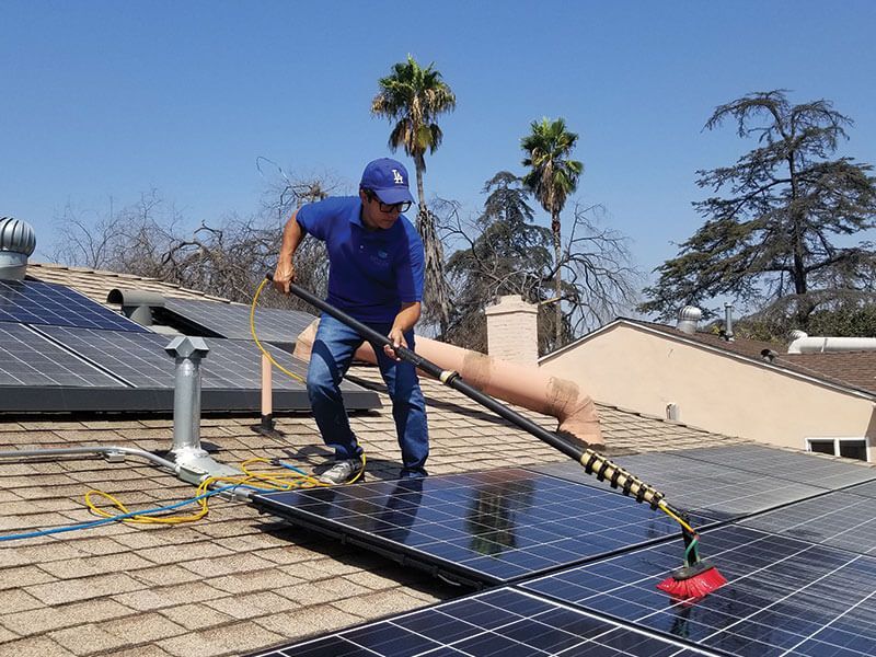 A person in a blue uniform cleaning solar panels on a shingled roof with a long-handled brush on a sunny day.