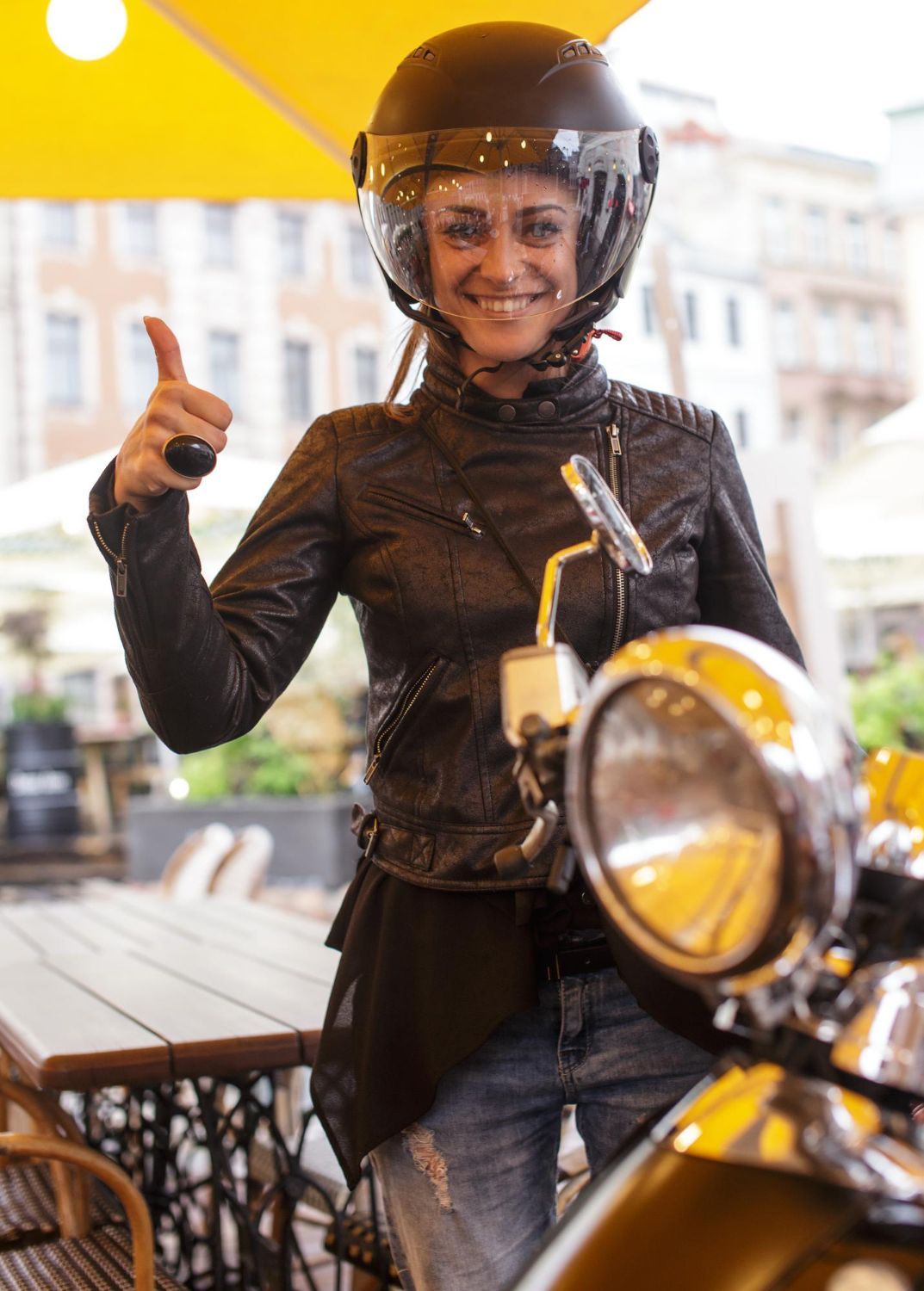 Woman in helmet and leather jacket, giving thumbs up near a scooter.