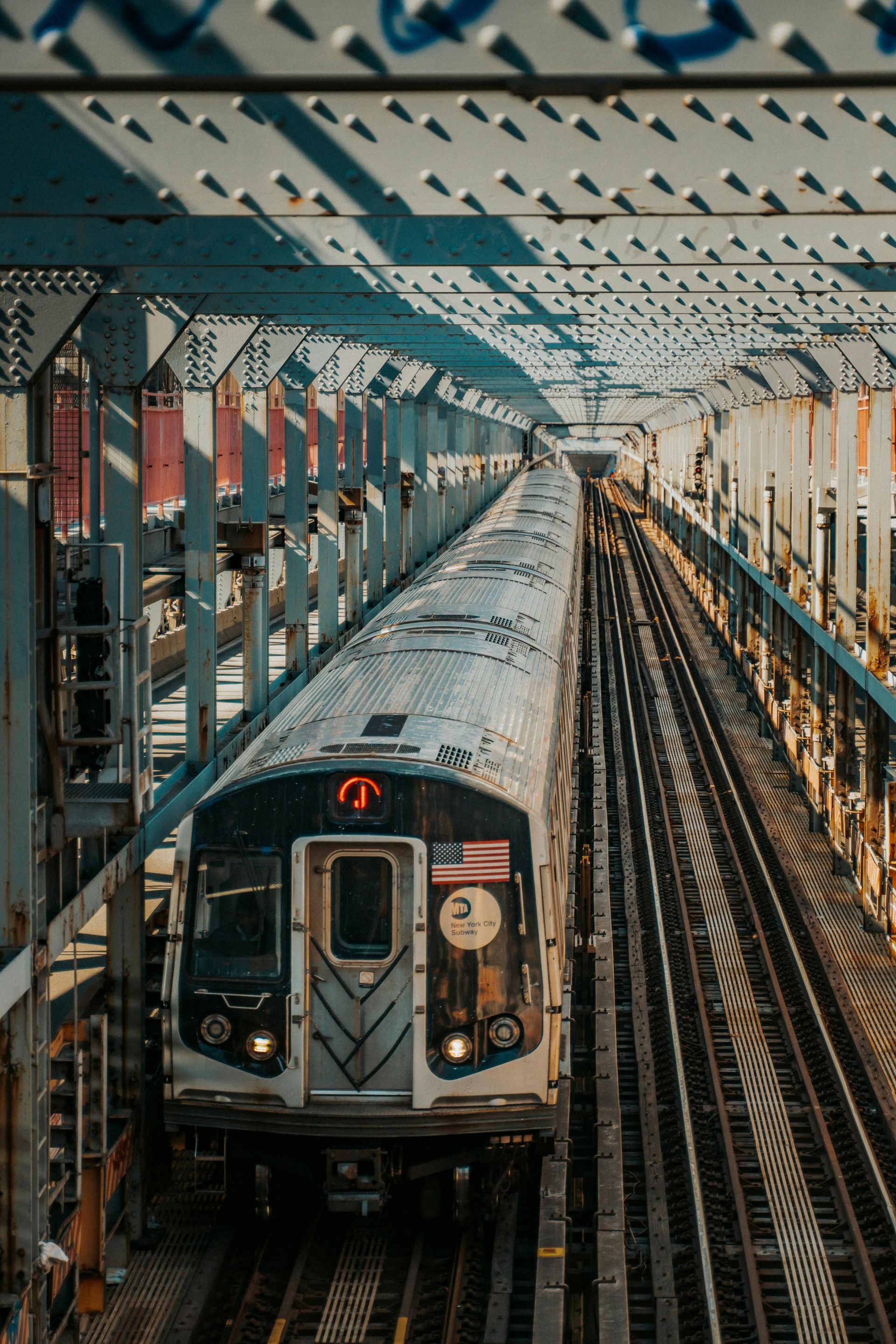 NYC subway train (2) on elevated tracks, passing under a metal bridge structure.