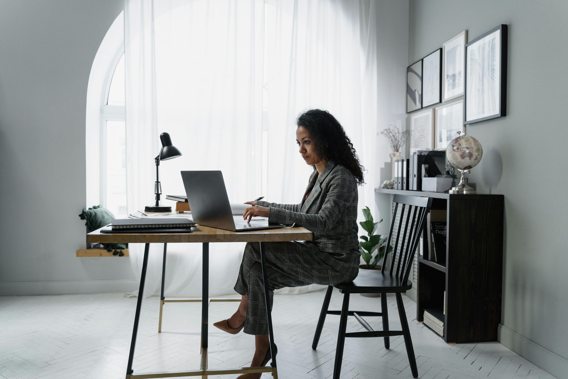 Woman in business suit works on laptop at desk by window.