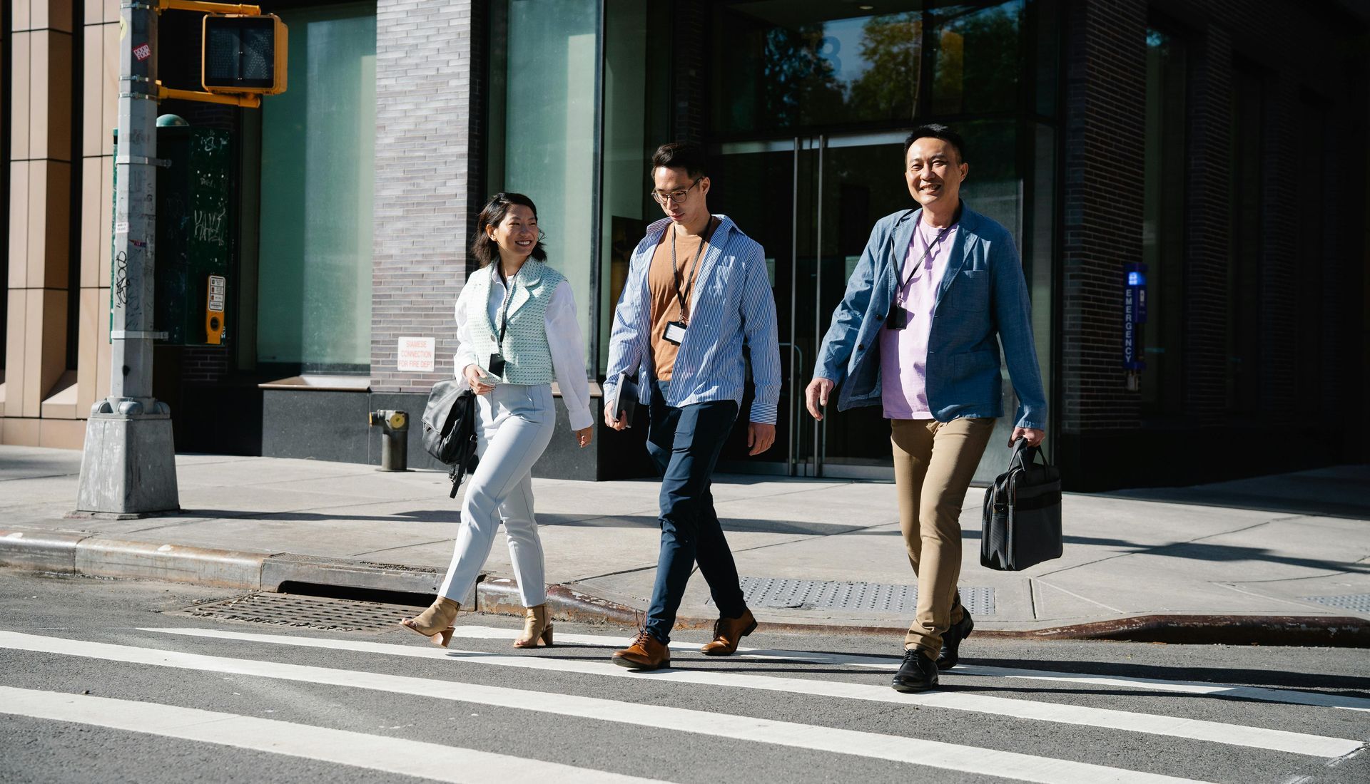 Three people crossing a street. Woman in white, two men in blue and tan, sunny day, urban setting.
