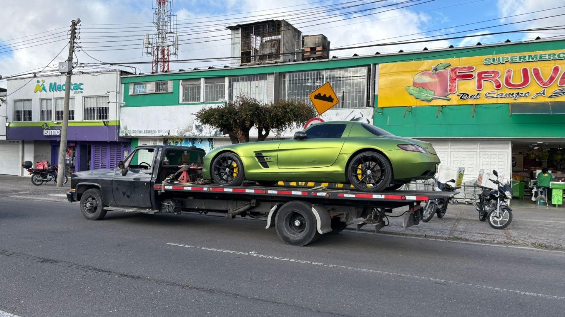 Un coche deportivo verde en una grúa pasando frente a unas tiendas en una calle de la ciudad