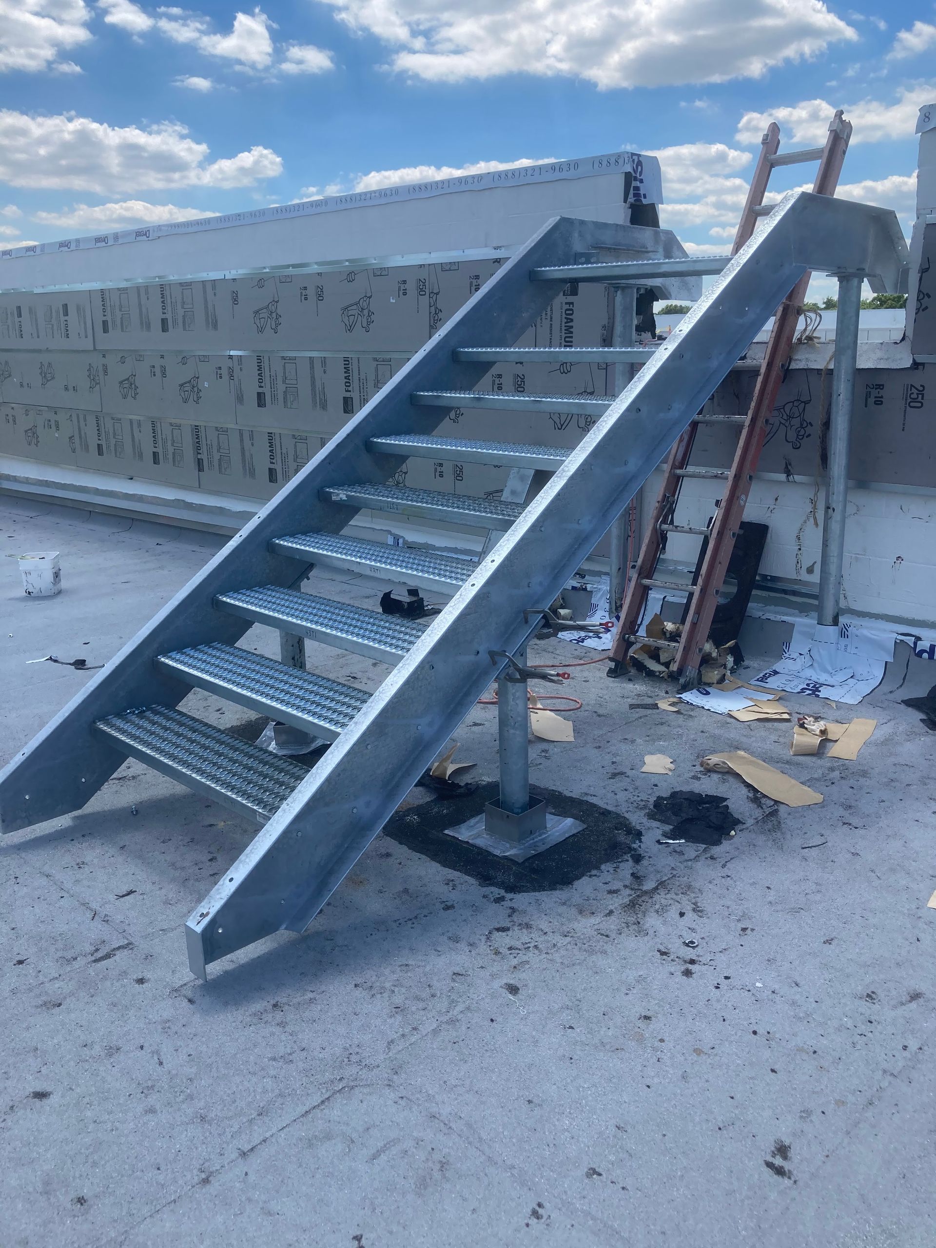 Metal rooftop access stairs leading to a flat roof, with a ladder leaning nearby against a wall.