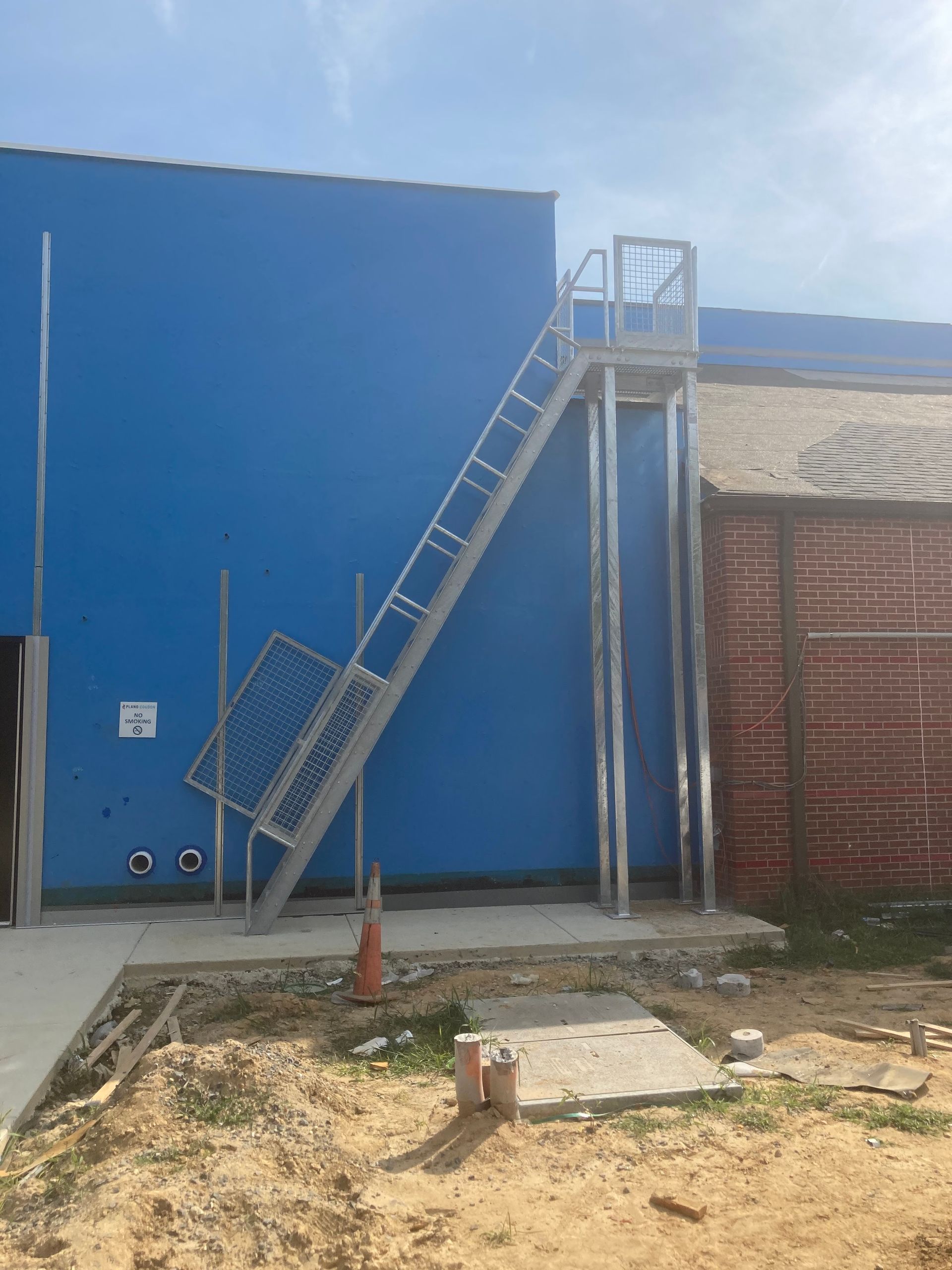 Metal ladder attached to a blue building leads to a platform with a railing. Orange cone and concrete.