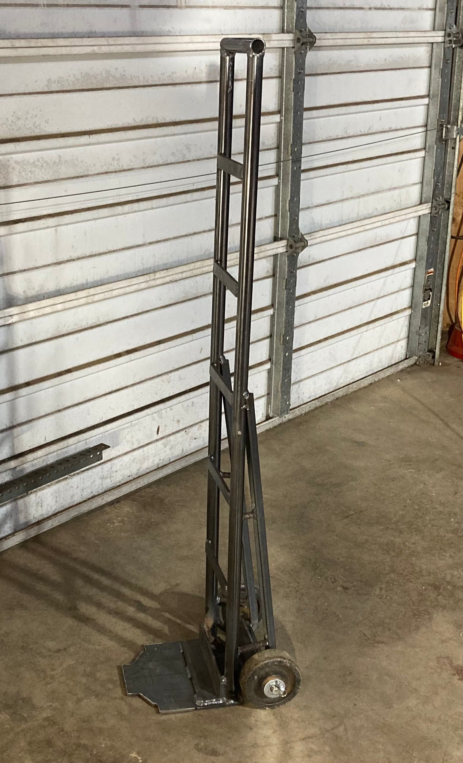 Metal hand truck with a narrow frame and wheels, positioned in front of a white garage door.