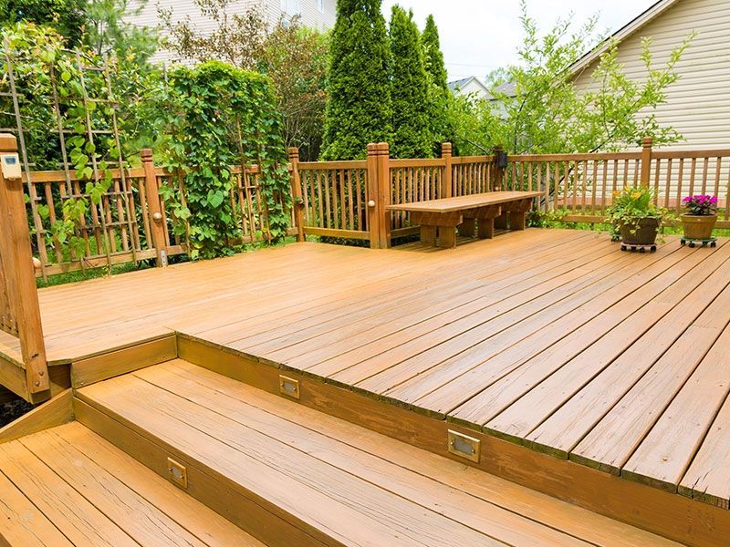 Wooden deck with stairs, built-in bench, and potted flowers. Green foliage surrounds the space.