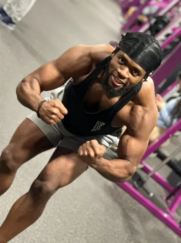 Muscular person flexing in a gym, wearing a black tank top, white shorts, and black cap.