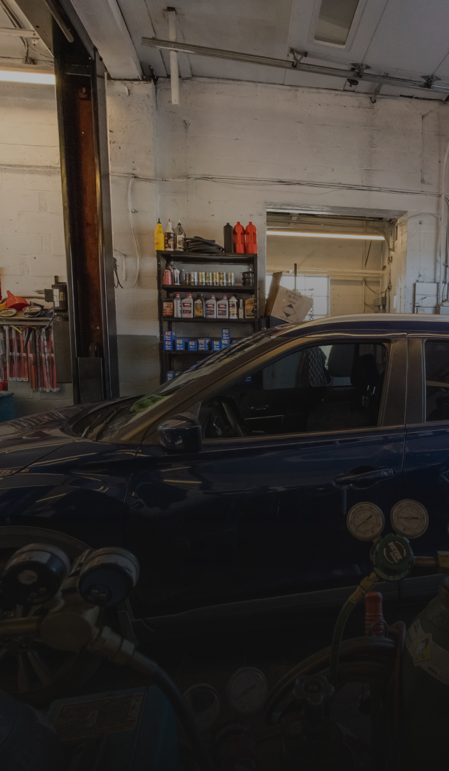 Dark blue car parked inside a garage with shelves and tools against a white wall. | Expert Auto Service & Licensing
