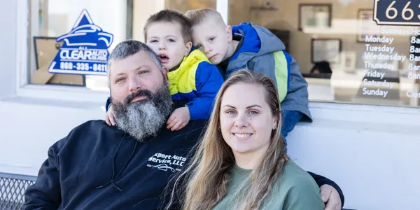 Family posing outside a storefront, seated and smiling, with a blue sign in the background | Expert Auto Service & Licensing