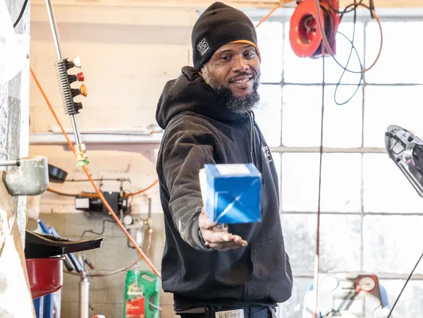 Person in a garage holds out a blue box, smiling, with tools and a window in the background | Expert Auto Service & Licensing