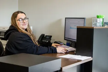 Woman at an office desk with a computer, phone, and papers, looking toward the camera. | Expert Auto Service & Licensing