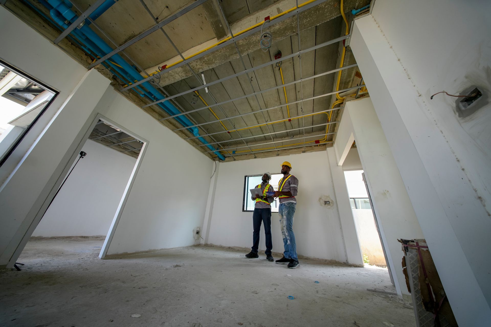 Two construction workers in hard hats inspecting a partially finished room with exposed ceiling wiring.