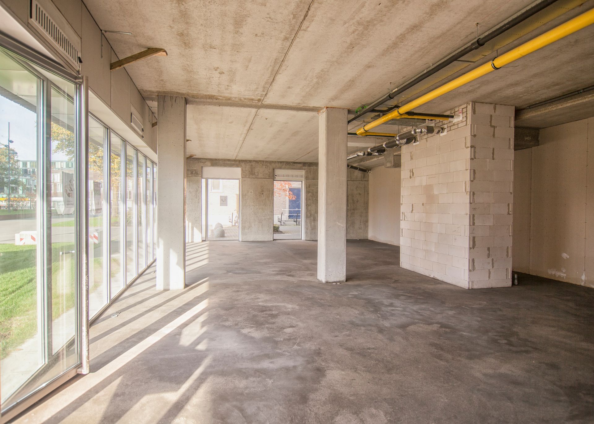 Empty unfinished concrete room with large windows and exposed yellow pipes.