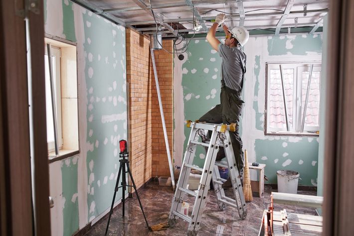 Person standing on a ladder during interior renovation, working on ceiling framing in a room with green drywall.