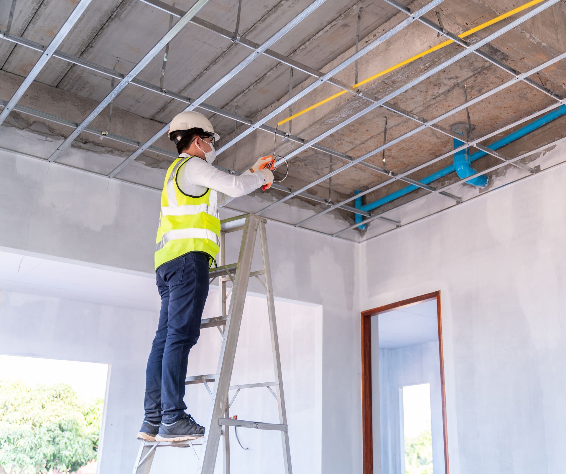 Worker in safety vest standing on a ladder, installing ceiling wiring in a room under construction.