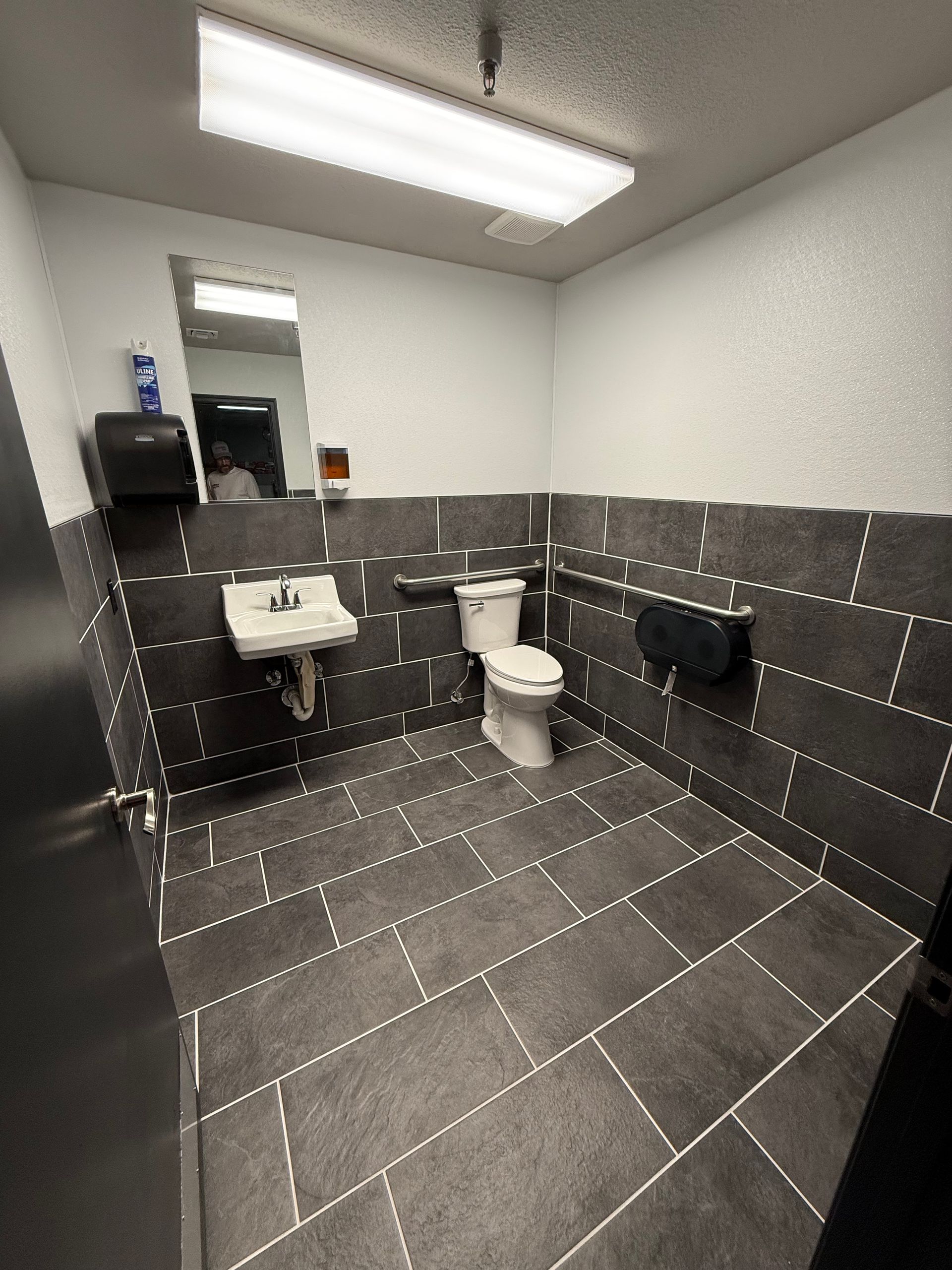 Small tiled bathroom with toilet, sink, mirror, and grab bars under fluorescent light.
