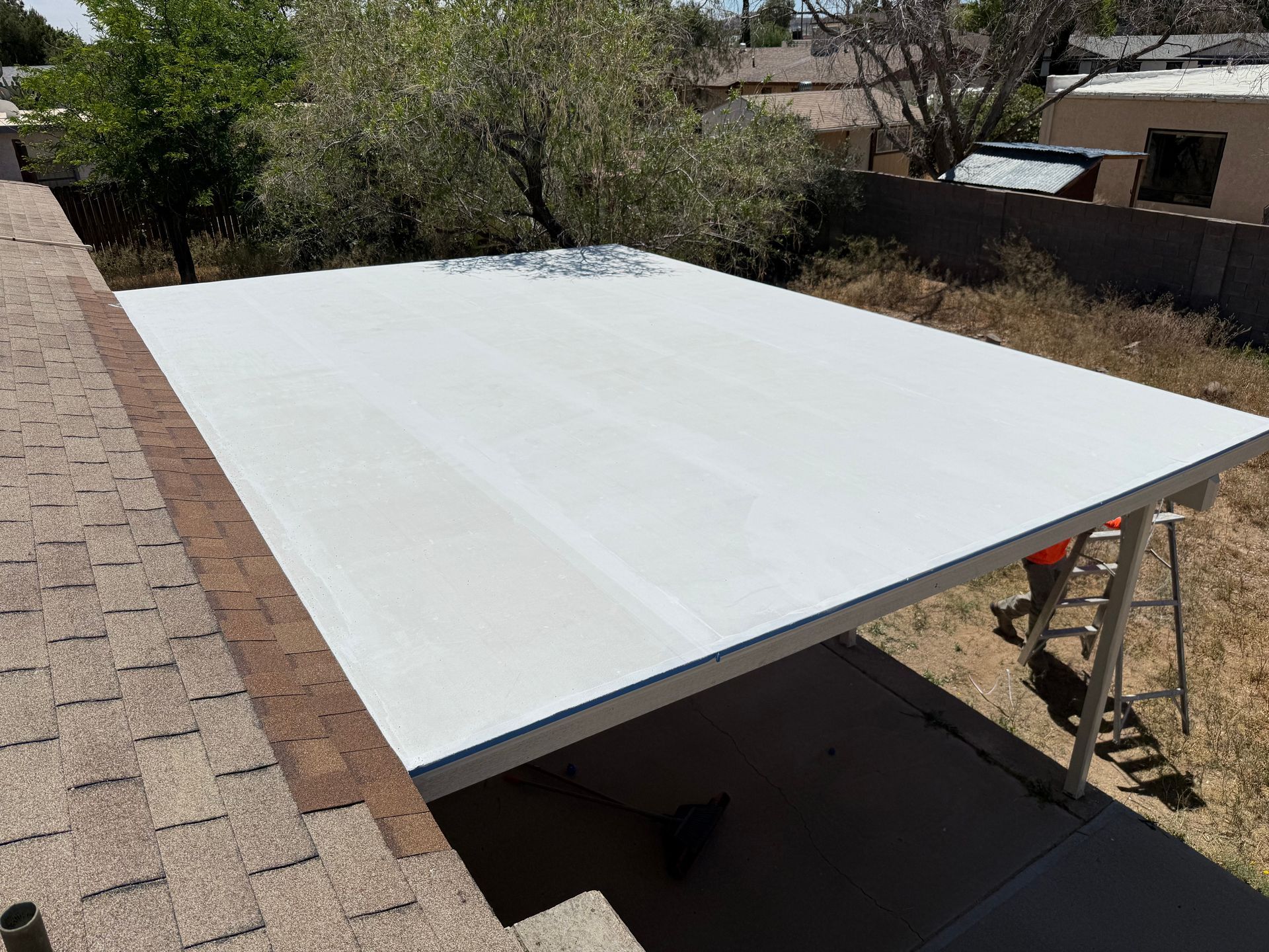 Large white roof panel on a raised outdoor structure in a dry yard, with trees and a brick wall nearby.