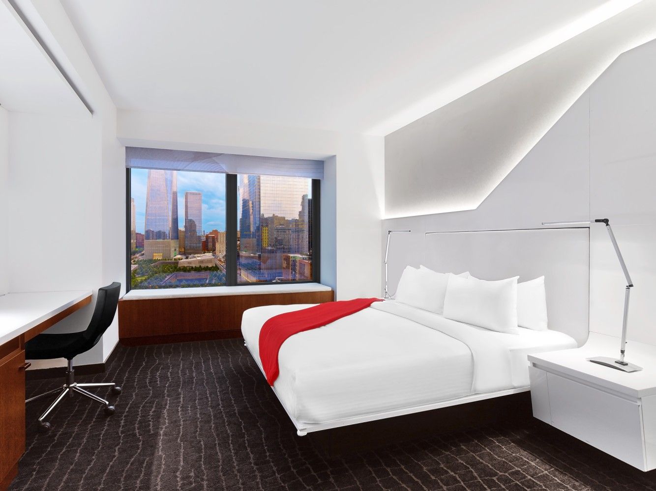 Modern hotel room with white bed, red throw, desk, and large city-view window