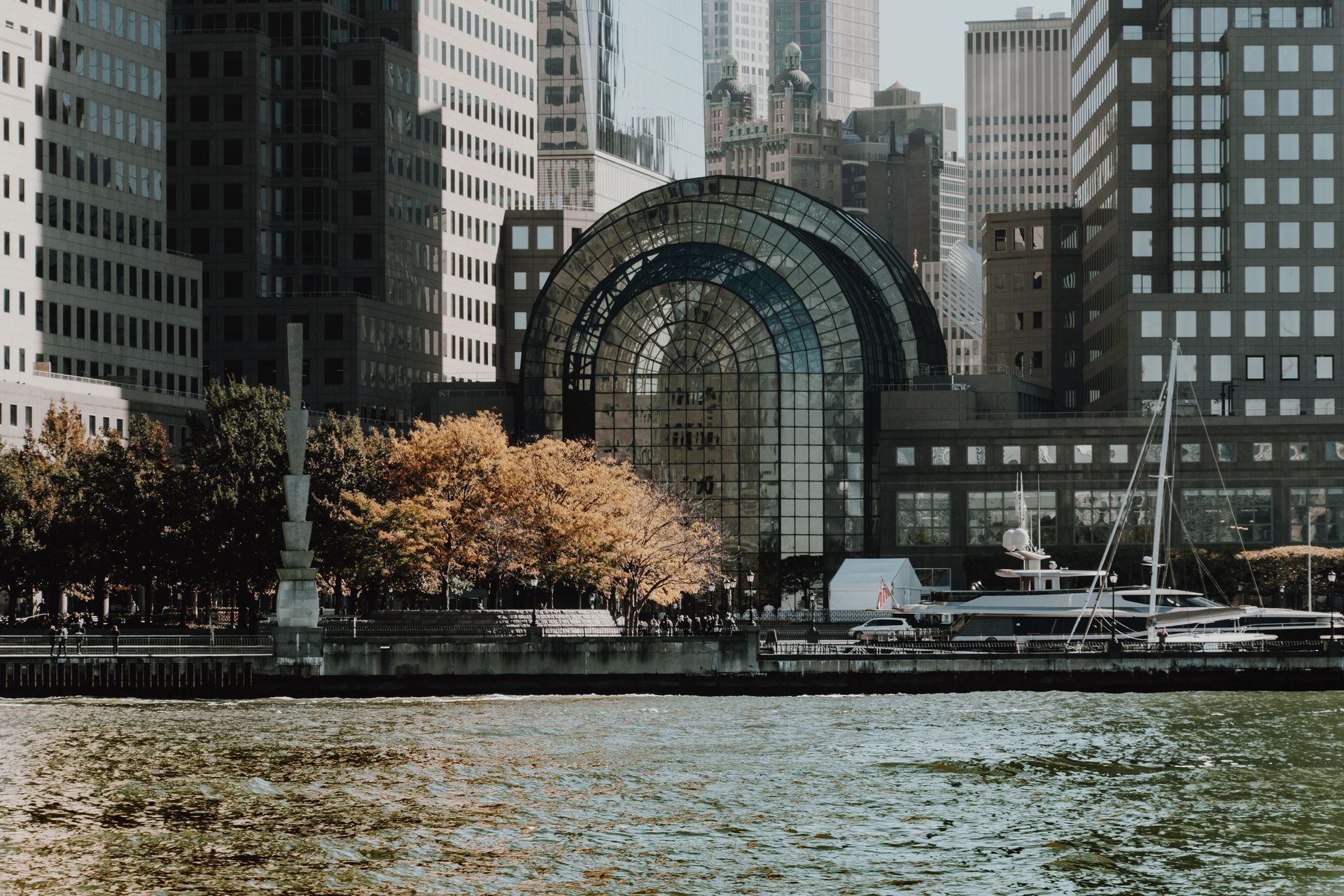 City waterfront with a large arched glass building, trees, and skyscrapers beyond the water.