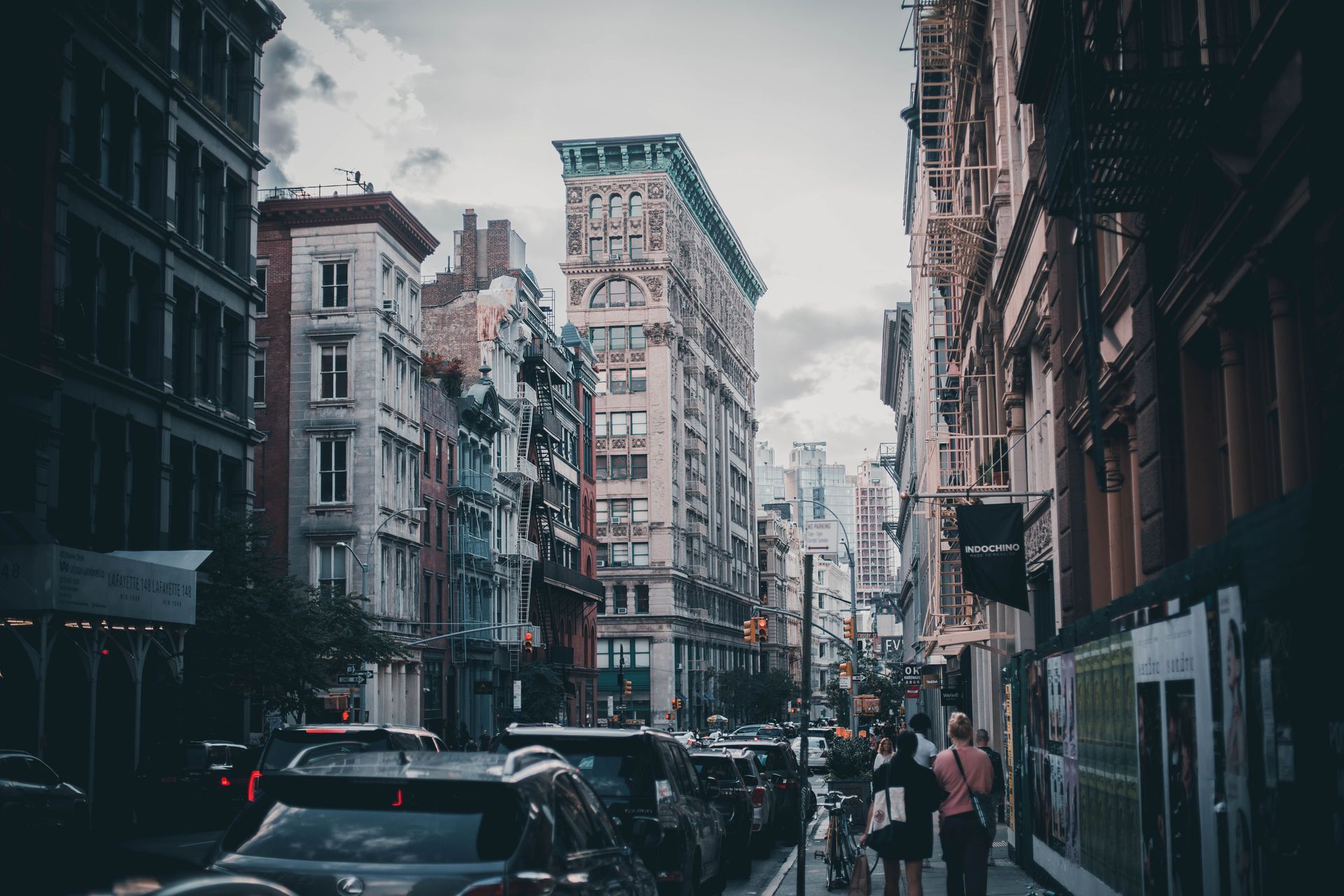 Narrow city street lined with tall buildings, parked cars, and pedestrians under a cloudy sky.