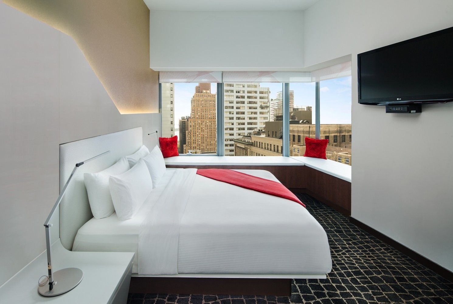 Modern hotel room with white bed, window seat, city view, and wall-mounted TV