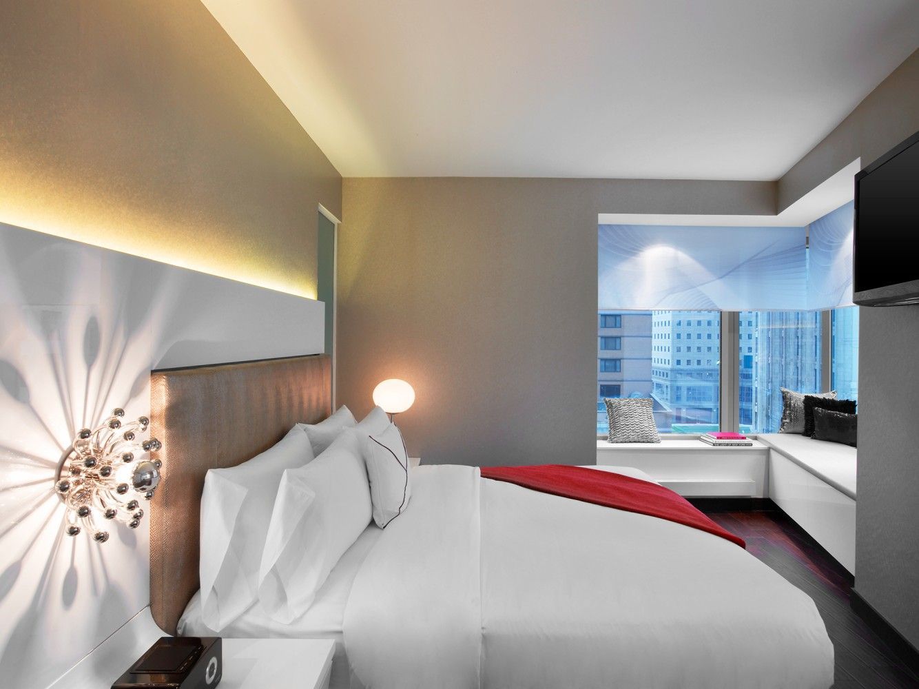 Modern hotel bedroom with king bed, city view window, and red accent blanket