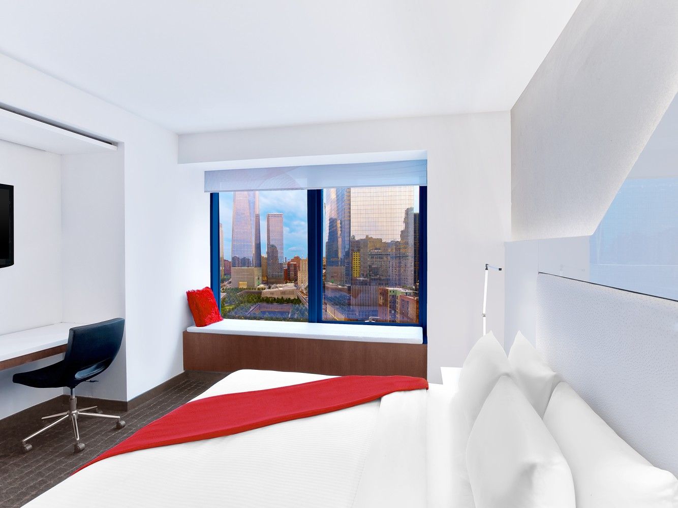 Modern hotel room with large city-view window, white bed, red rug, and desk nook