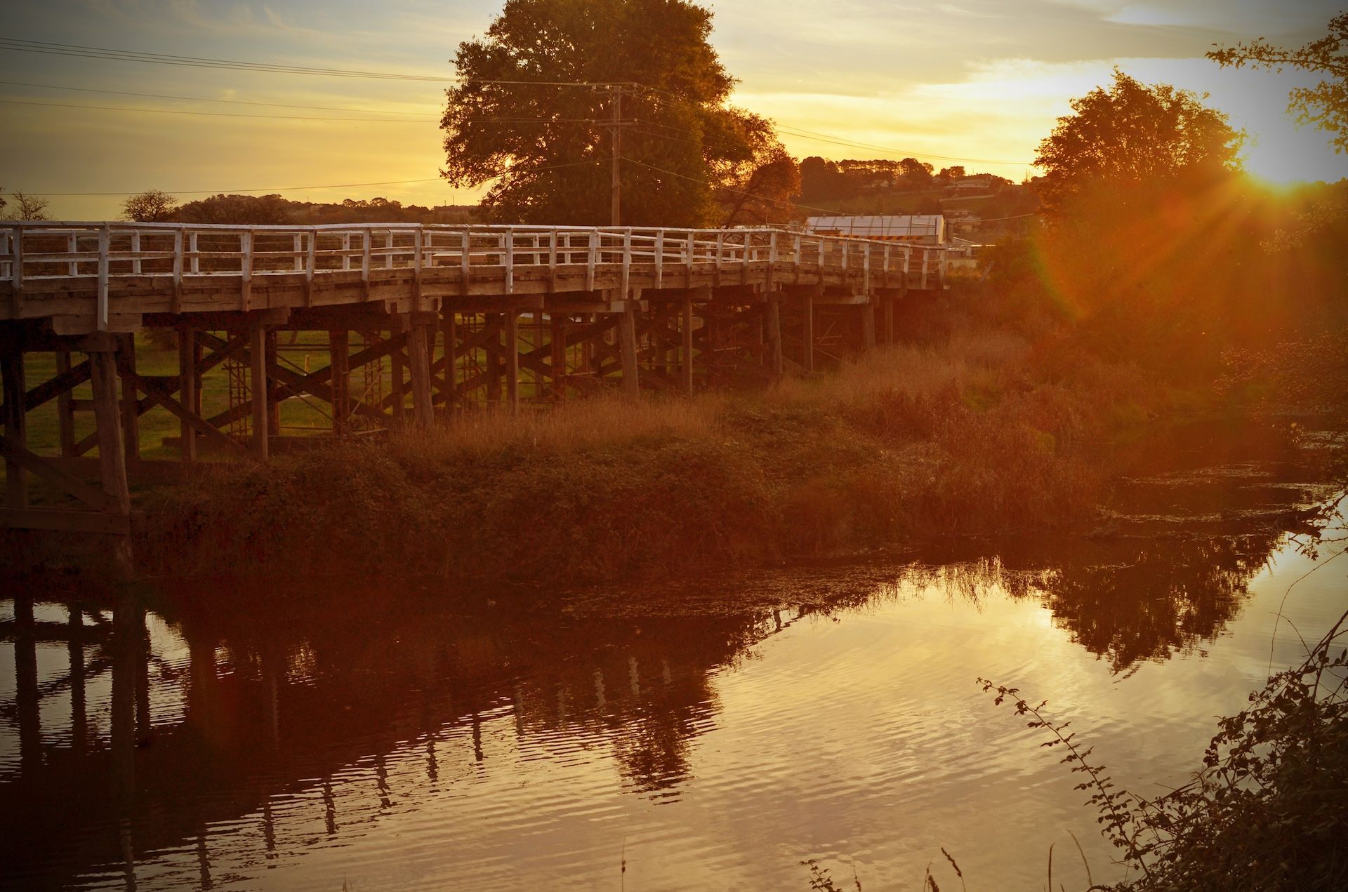 Wooden bridge over water at sunset, reflecting in the water. Sunlight bursts from behind trees — JSF Interiors Goulburn in Goulburn, NSW