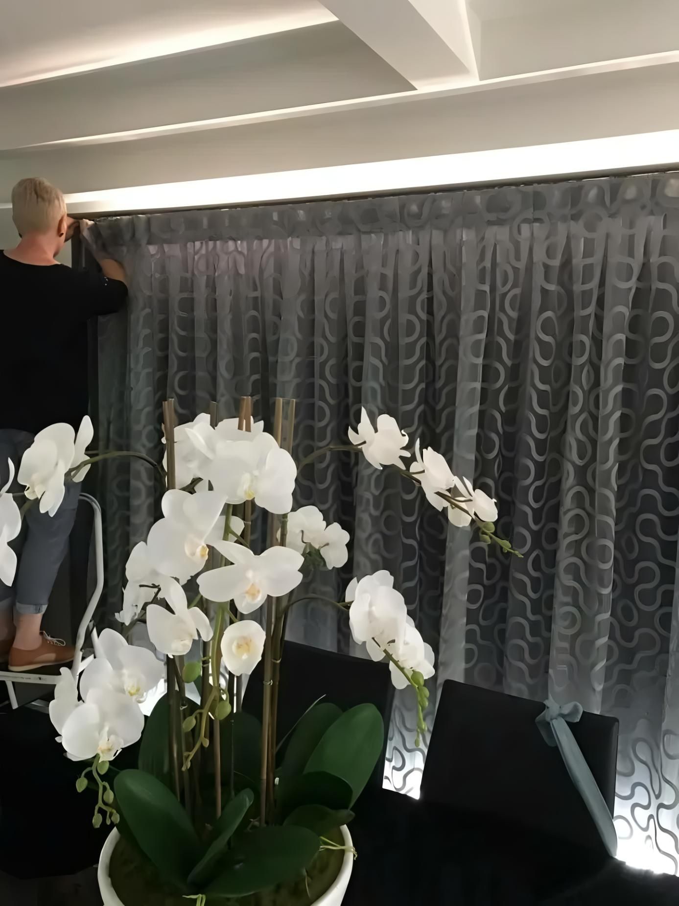 Person adjusting gray patterned curtains, standing on a step ladder. White orchids in foreground, next to a black table 